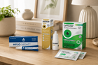 A group of eye care products arranged on a wooden tabletop in a softly lit room: a blue Hylo Night tube and box, a yellow Hycosan Shell box, a white Optase box with green graphics, and two Optase single-use packets in front. A lamp, framed art, books, and a plant are blurred in the background.