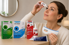 A woman applies Optase Night Eye drops to her eye while holding a Optase Night contact lens case. Four product boxes and tubes for Optase and Hycosan are lined up on a wooden table in a warm, softly lit room.