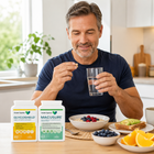 A man in a navy blue t-shirt sits at a kitchen table, holding a pill and a glass of water. In the foreground, two white supplement bottles labeled 'Kubera Organics' (GlycoShield and Macusure) are displayed next to bowls of fruit and oatmeal.