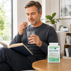 A man sitting in a chair by a window reads a book and holds a glass of water, with a bottle of Double Express MACUSURE supplements placed on the wooden table in the foreground.