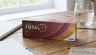 A box of Daies Total 1 contact lenses by Alcon sits on a wooden table next to a glass of water, with notebooks in the background.