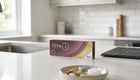 A box of Dairi Total 1 contact lenses by Alcon sits on a white kitchen countertop, with a blurred kitchen background and a small bowl of contact lens solution capsules in the foreground.