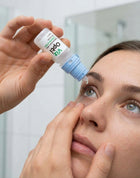 A close-up lifestyle shot of a woman applying eye drops from a small white and blue bottle held by another hand, with a bathroom background.