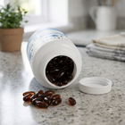 An open white supplement bottle lying on its side on a speckled granite countertop, with dark brown softgel capsules spilling out. The white lid sits beside the bottle, and a blurred kitchen background with a potted plant and folded towels is visible.
