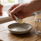 A hand holds a small white spoon containing a gold-colored softgel capsule over a shallow speckled ceramic dish on a wooden table, with a glass of water nearby.