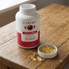 A white supplement bottle labeled VITEYES 2 Zinc-Free stands on a wooden table next to an open cap filled with yellow softgel capsules.