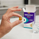 A hand holds a small amber-yellow eye drop capsule in front of a blurred Macusave Gold eye health box and a glass of water on a light countertop.