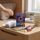 VisionaCe MAX eye drops packaging box standing on a wooden table, with blister packs of yellow and red capsules and a hand reaching in from the left in a bright indoor setting.