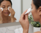 A woman in a bathroom uses a white facial cleansing pad to wipe her face, with her reflection visible in the mirror behind her. She is also using cotton pads on her cheeks.