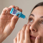 A close-up lifestyle shot of a woman applying eye drops from a small blue and white bottle labeled 'LUMIRES' to her eye.