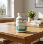 A white bottle of Glaucomide dietary supplement by Ocular Express sits on a wooden table in a bright, modern room with a window, books, and a small plant in the background.