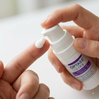A close-up of a person's hand dispensing white eye drops onto another finger, with the Optase Life sensitive eye daily drop bottle held in the other hand.