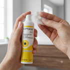 A person holds a bottle of OPTASE Eyelid Cleansing Gel in one hand and applies gel with a cotton pad in the other, set against a softly blurred indoor background.