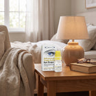 A bottle of eye drops and its packaging box sit on a wooden bedside table in a softly lit bedroom. The box reads 'Advanced Triple Action Eye Drops' and shows a white-and-blue design with an eye graphic.