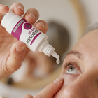 A close-up lifestyle shot of a person applying a drop of eye drops from a white and purple bottle held by another hand.