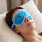 A woman lying down wearing a blue Optase Cooling Mask eye mask over her eyes, with a beige pillow and neutral background.
