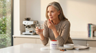 A woman in a beige sweater sits at a kitchen counter holding a glass of water and a red pill, with a jar of CalibreMind supplement and other small items on the counter in a bright, modern kitchen.