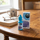 A bottle of Blink Intensive soothing eye drops for cats and dogs stands on a wooden table next to its product box, with a blurred book, glasses, and mug in the background.