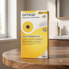 A box of Optase Manuka Oil Eyelid Wipes stands on a round wooden table in a softly lit indoor setting. The packaging is predominantly yellow and white with a large eye graphic and text highlighting cleansing, soothing, and hydrating benefits.