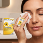 A woman with closed eyes wipes her face with a white paper towel while holding an OPTASE Manga Oil Eyelid wipes packet. The yellow and white product box is visible in the background on a bathroom counter.