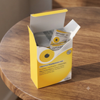 A yellow and white SCOPE Optase mouth oil pouch box sits open on a round wooden table, with two individual Optase pouches visible inside.