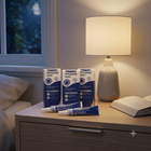 Three boxes and two tubes of Hycosan Night eye drops are arranged on a wooden bedside table next to a lit table lamp, with a bed and window in the background.