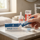 A hand holds a blue and white tube of HYLO NIGHT eye drops next to its packaging on a bathroom counter, with other HYLO products and contact lens cases in the background.
