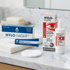 Ursapharm Hylo Night lubricant kit on a white marble bathroom counter, including a blue and white tube, a blue and white box, a white bottle, and cotton pads.
