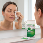 A woman in a bathroom applies a wet wipe to her eye while looking into a mirror. In the foreground on the counter are Optase eye wipes packaging boxes and a single packet.