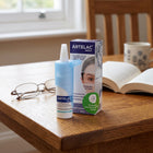 Artelac Night Moisturizing Eye Drops 10ml bottle and packaging box standing on a wooden table next to eyeglasses and an open book, with a blurred indoor background.