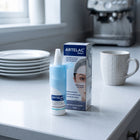 Artelac Night Moisturising Eye Drops bottle and packaging box standing on a light countertop in a kitchen setting, with plates, a mug, and a coffee machine blurred in the background.