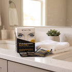 A box of Eyecure biodegradable eyelid wipes sits on a bathroom counter next to two individual packet wipes, with a white towel and a small potted succulent in the background.