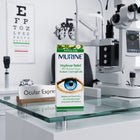 A box of Murine Hayfever Relief 2% Mydriatic eye drops stands on a glass display shelf in an eye care clinic, with a验光师 exam light, slit lamp, and eye chart visible in the background.