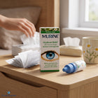 A box and bottle of Murine Hayfever Relief 2% eye drops sit on a wooden table in a softly lit room, with a hand holding tissue paper in the background.