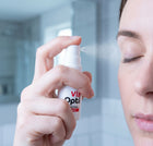 A close-up of a person holding a ViziOpti eye function spray bottle in front of their face, with the spray mist visible. The background is a softly blurred bathroom setting.