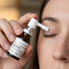 A close-up of a person applying eyelid oil from a small amber bottle with a white dropper applicator to their closed eye. The bottle label reads 'Advanced Comfort Eyelid Oil' and shows the brand 'Gyvena'.