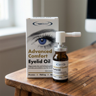 Eyedoctor Advanced Comfort Eyelid Oil shown with its retail box and a small amber glass vial with a white dropper cap, placed on a wooden table in a softly lit indoor setting.