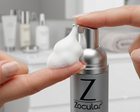 A close-up of a silver and black Zocular foaming bottle with white foam being dispensed onto a finger, set against a blurred bathroom vanity background.