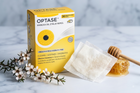 Optase Manuka Oil Eyelid Wipes shown with a yellow and white product box, a single unfolded white wipe, a wooden honey dipper with honey, and small white flowers on a light marble surface.