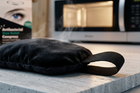 A black fabric eye bag cooling pad with a loop handle sits on a light gray stone countertop, with visible steam rising from it. A microwave is blurred in the background, and a product box for an antibacterial eye bag is partially visible on the left.