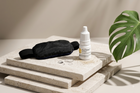 A small white bottle of EvoLabs Lens Care liquid for contact lens care stands on stacked beige stone slabs beside a black eye mask, with two contact lenses and a green monstera leaf in a warm studio setting.