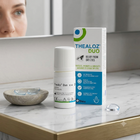 Thealoz Duo Relief from Dry Eyes eye drops bottle and packaging box on a marble bathroom counter with a person's face blurred in the mirror background.