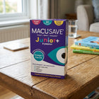 A box of MACUSAVE Junior+ 3 years+ supplement placed on a wooden table in a home setting, with books and a glass of water blurred in the background.