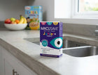 A box of Macusave Junior+ 3 years+ liquid drops stands on a light gray kitchen countertop near a sink, with a blurred cereal box and fruit bowl in the background.