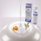 A clear glass wine glass base with a stem, shown on a white surface with honeycomb, a small sprig of green herb, and clear beads in the foreground. In the blurred background are two Optase Life products, a box and a bottle, on a clean white studio background.