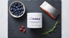 A white ReMind supplement jar sits on a dark slate board, surrounded by blueberries, a salmon fillet, two red capsules, and a rosemary sprig.