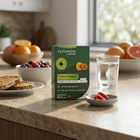A box of eyetamins by Optivue Vision Support dietary supplements sits on a kitchen counter next to a glass of water and a small dish of red capsules, with breakfast foods and a window in the blurred background.