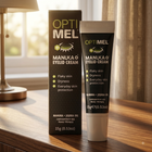 OPTIMEL Manuka Eyelid Cream shown as a black squeeze tube next to its matching black and gold outer box, placed on a wooden table in a warm, sunlit room.