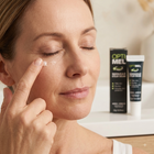 A woman applies a small amount of cream to her cheek while standing in front of skincare packaging. The products behind her read Opti Mel and Manuka Daily Cream.