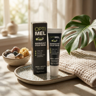 OPTI MEL Manuka Eyelid Cream shown with its black product box and matching squeeze tube on a ceramic tray, styled with a woven towel, a bowl of crystals, and a large green monstera leaf in a sunlit room.